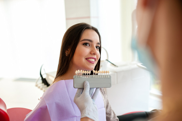 Dentist holding a shade guide next to a smiling female patient's teeth during dental veneer preparation.
